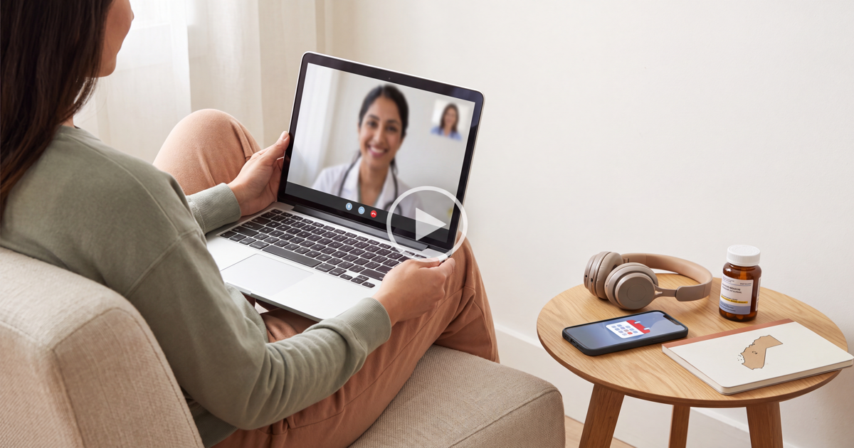 Woman sitting on a couch video-calling a clinician on her laptop, with headphones and a medication bottle on the side table beside her